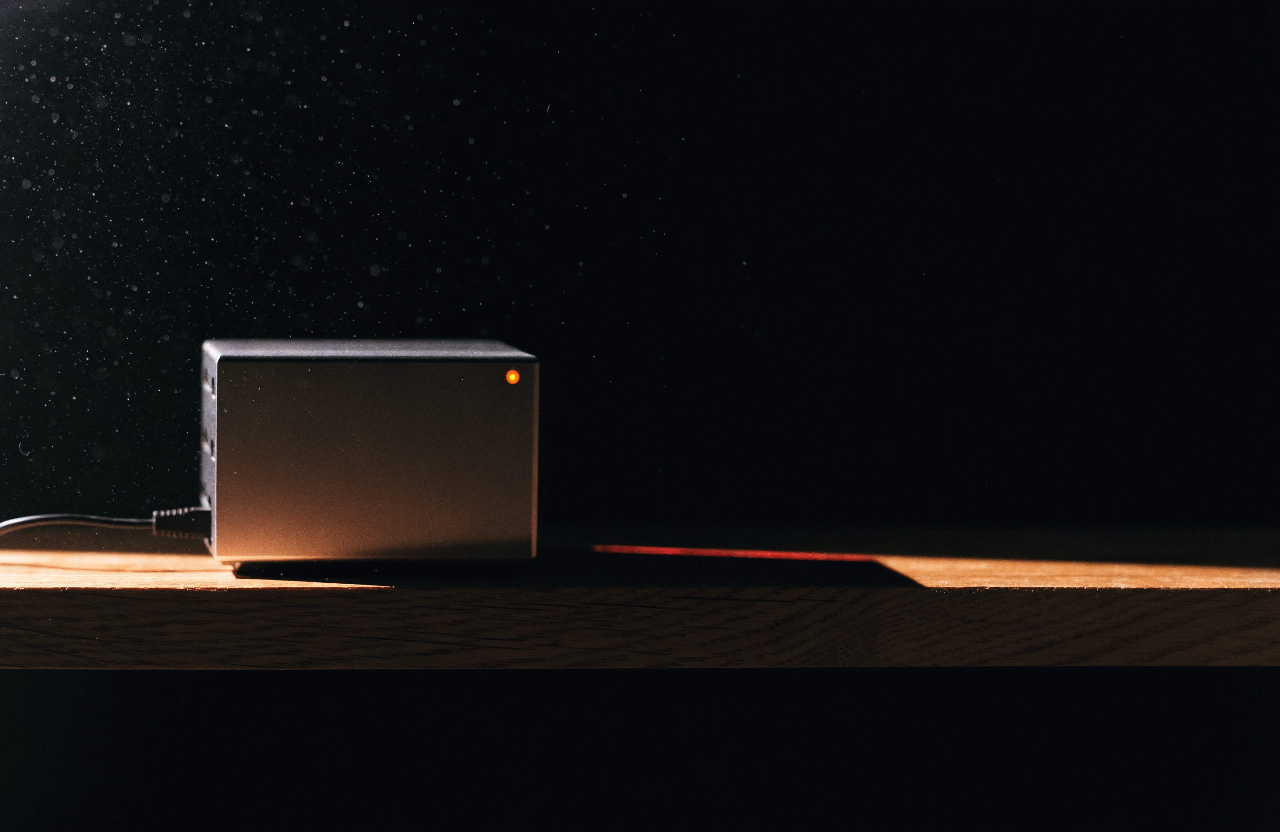 A small fanless aluminium mini-server on a bare wooden shelf in a dark room, one amber status light, a single ethernet cable disappearing into the dark, dust caught in a hard side light.